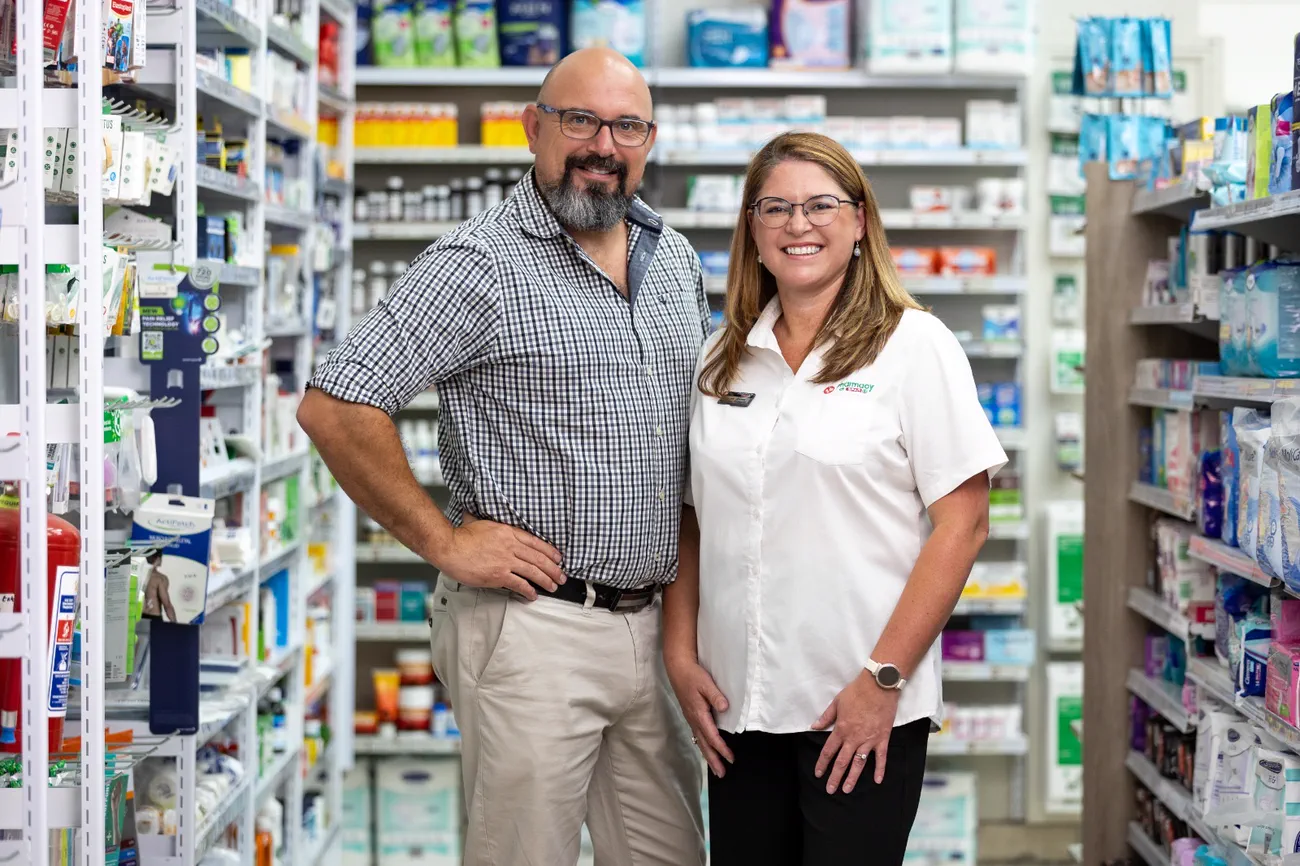 The owners of Albertyn Pharmacy standing together in an aisle of the shop, shelves stocked either side of them